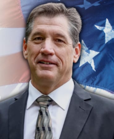 A man in a suit and tie smiles against a blurred American flag background.