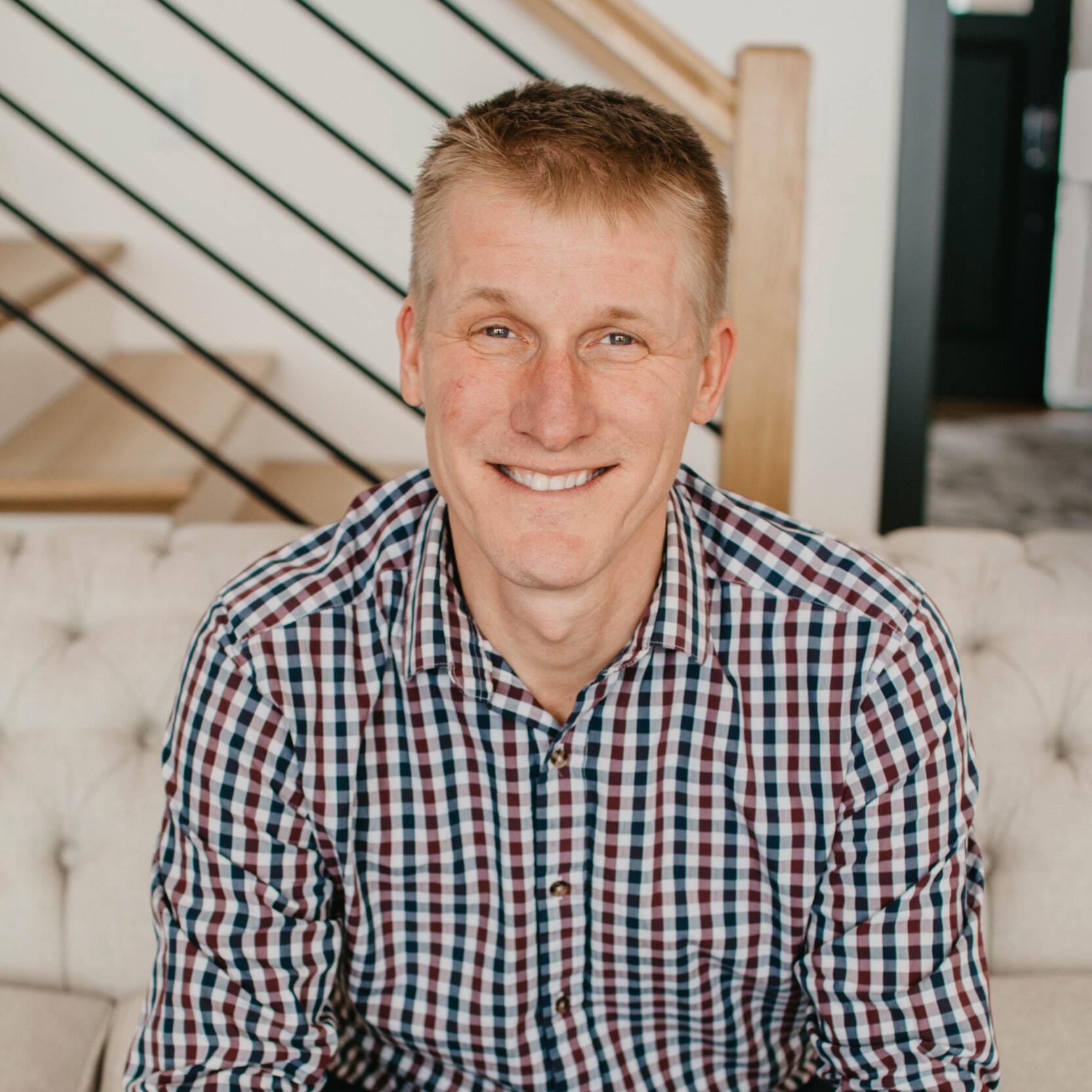 A smiling man in a plaid shirt sits on a couch, looking at the camera.