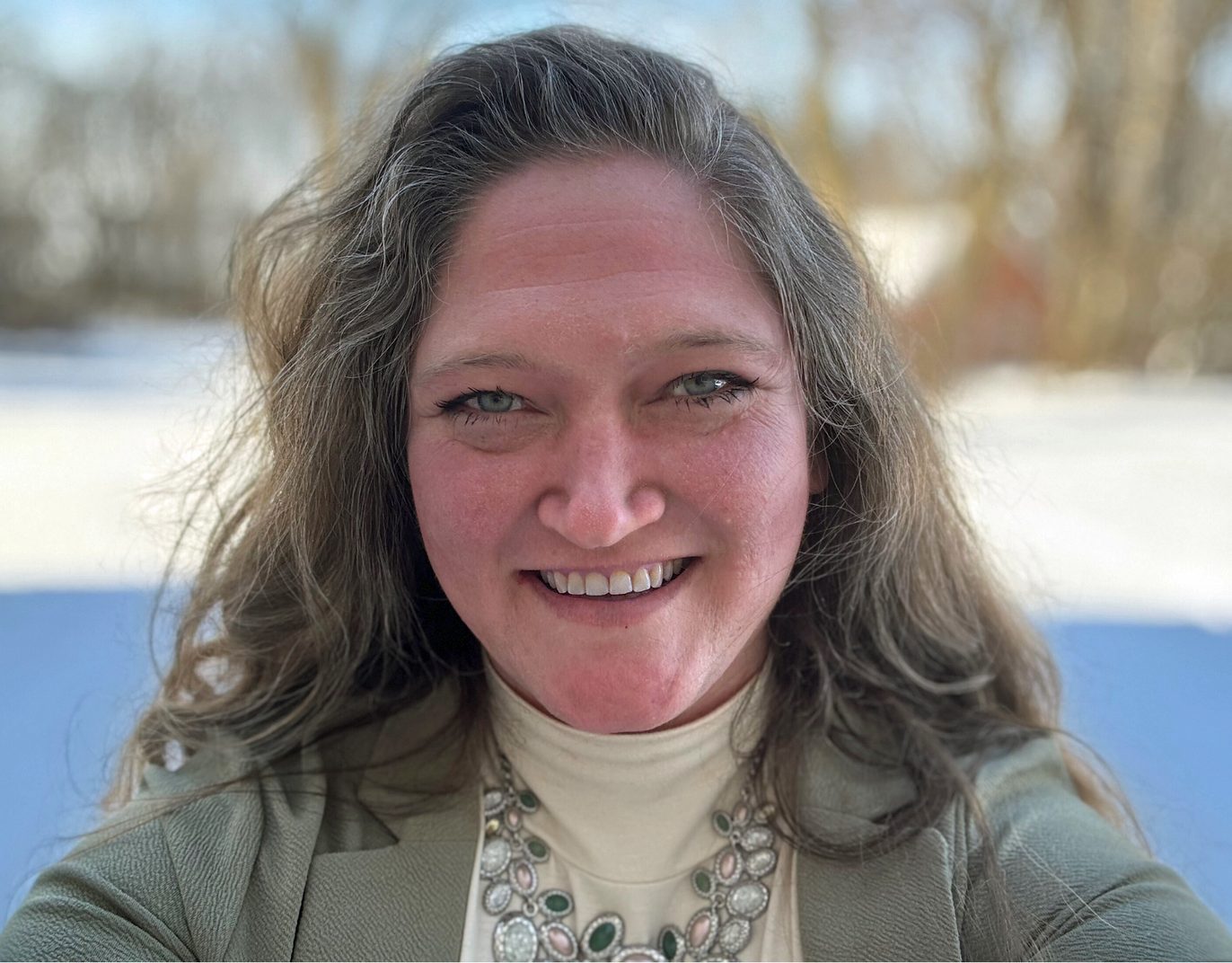 A smiling woman with long wavy hair and blue-green eyes wearing a necklace and green jacket in a snowy setting.