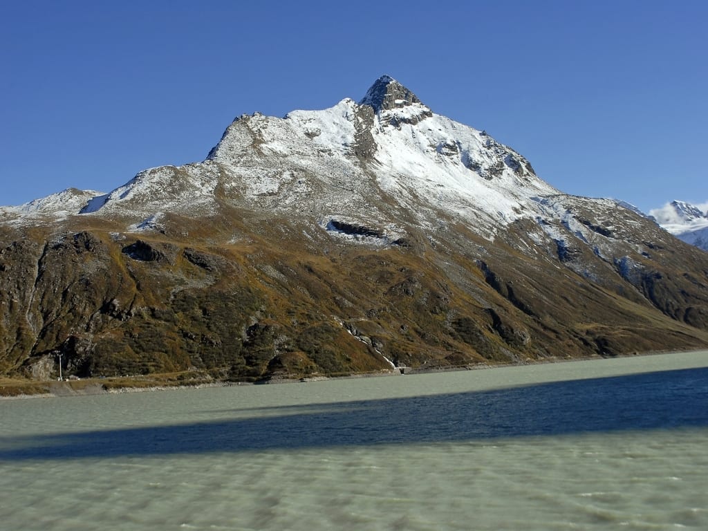A majestic snow-capped mountain rises above a serene lake under a clear blue sky, showing rugged terrain.