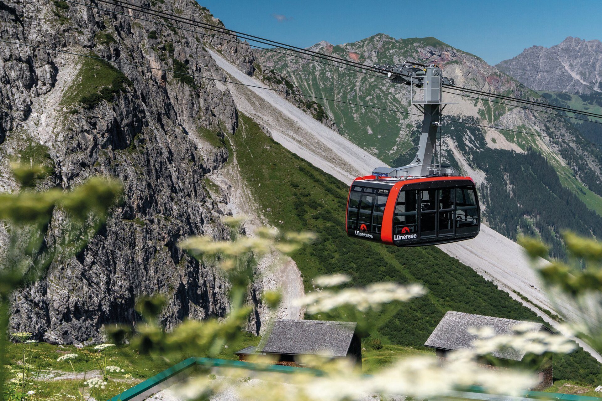 A red and black gondola car suspended on a cable over a rugged mountain landscape with green slopes and rocky peaks.