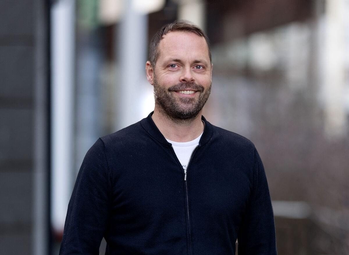 A smiling man with a beard and short hair, wearing a dark zip-up jacket, looks at the camera.