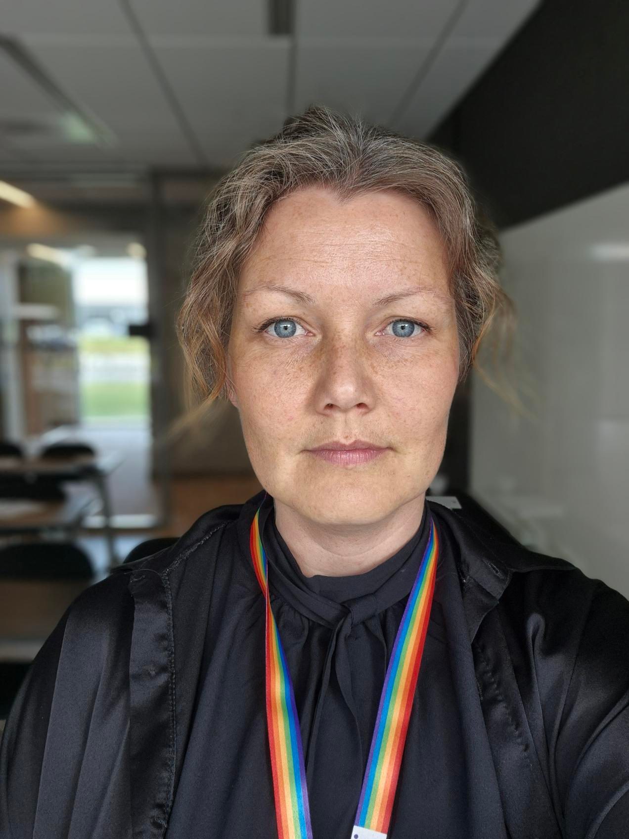 A woman with blue eyes, light brown hair, and a rainbow lanyard, looking directly at the camera.