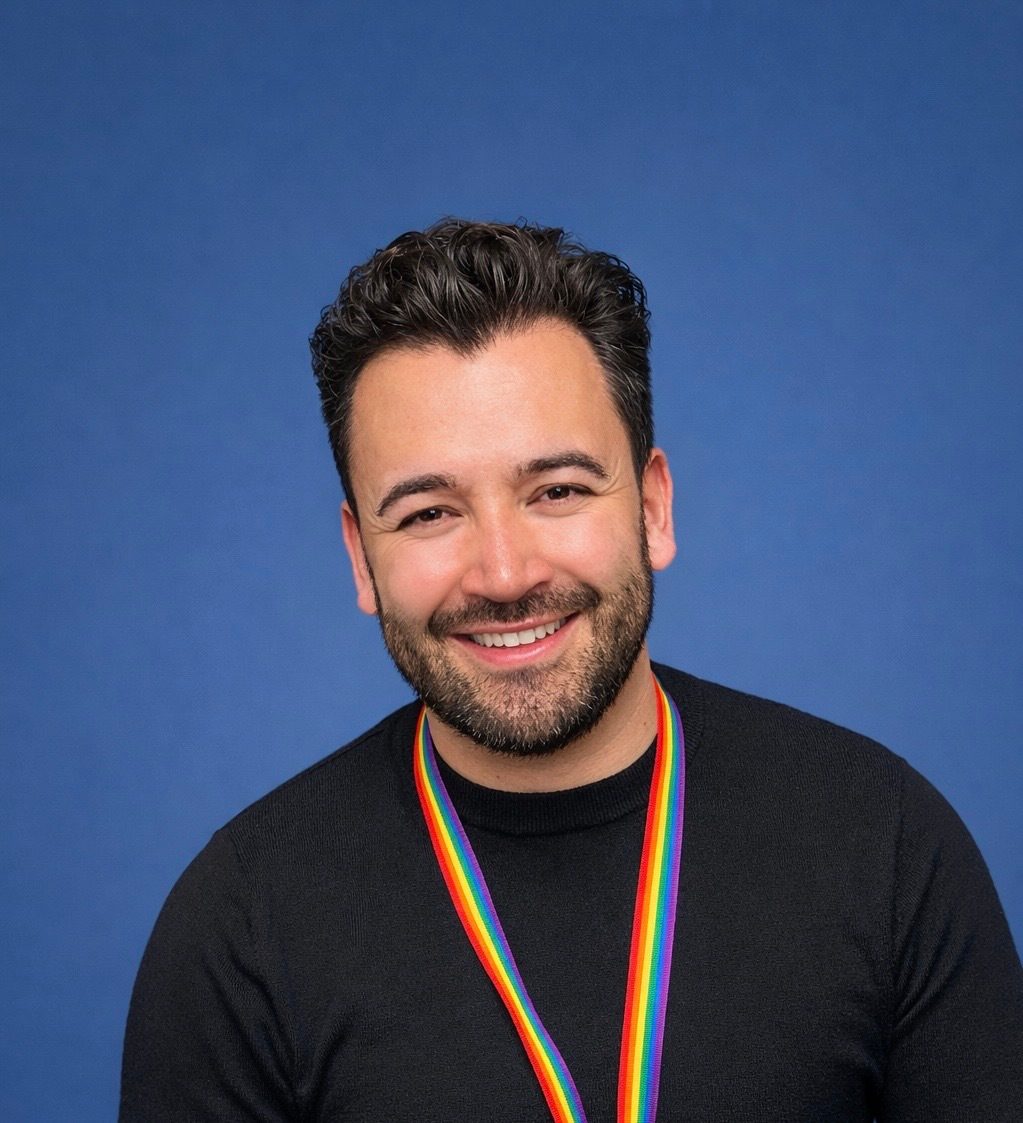 A man with a beard and curly hair smiles, wearing a black shirt and a rainbow lanyard against a blue background.