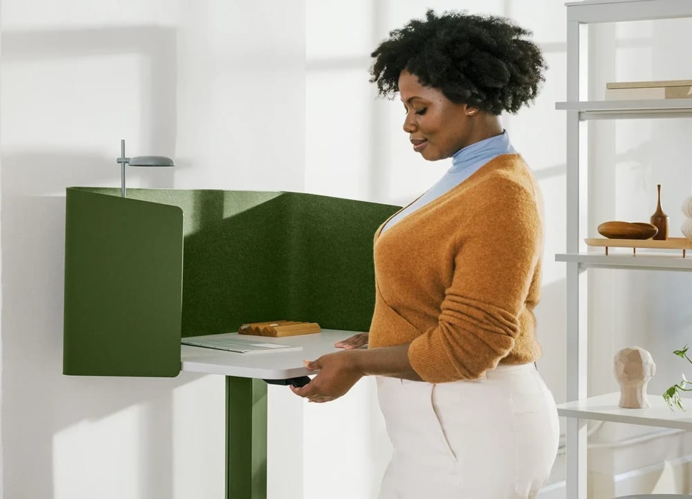 Woman adjusting a green felt standing desk workstation, with shelves on the right.