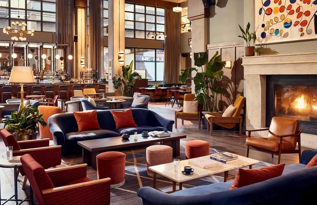 A warm and stylish hotel lounge with mid-century modern furniture, including navy blue sofas, leather armchairs, and orange accent pillows, arranged around a lit fireplace. The space features high ceilings, tall windows, and a well-stocked bar in the background.