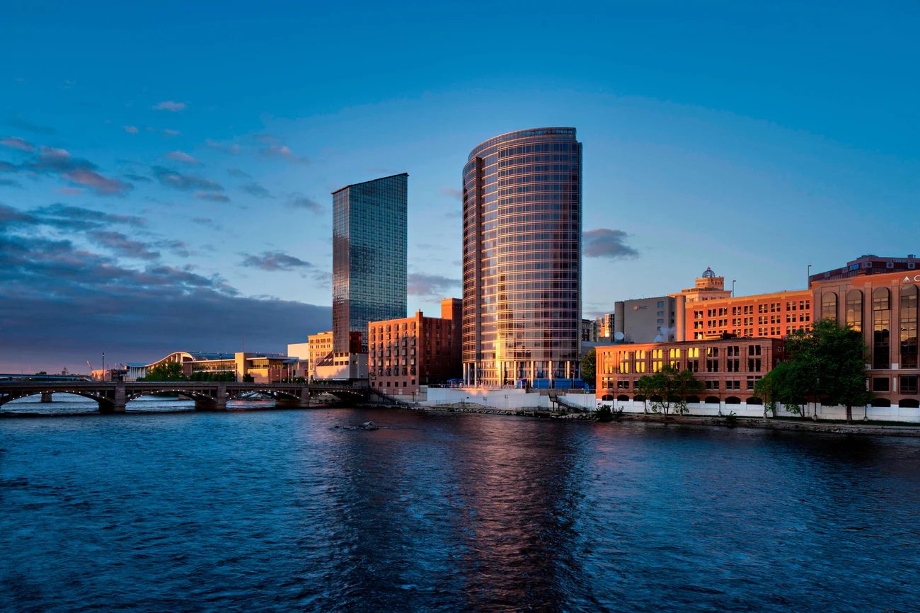 Exterior view of the JW Marriott Grand Rapids, a tall modern glass tower overlooking the Grand River. The curved façade reflects the sunset sky, with the city’s skyline visible around the riverfront.