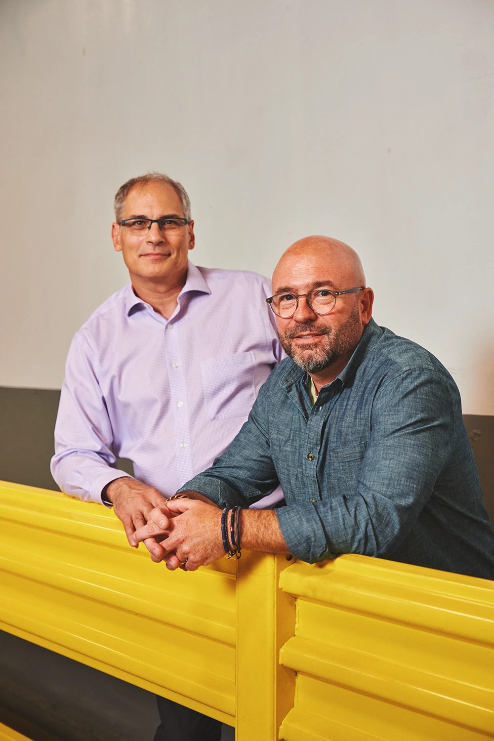Two men in shirts and glasses lean on a yellow barrier.