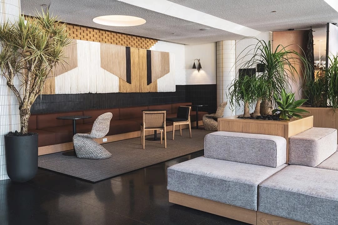 Modern boutique hotel lobby with light gray modular seating, woven lounge chairs, and natural wood accents. Large potted plants and a textured wall installation create a warm, contemporary atmosphere.