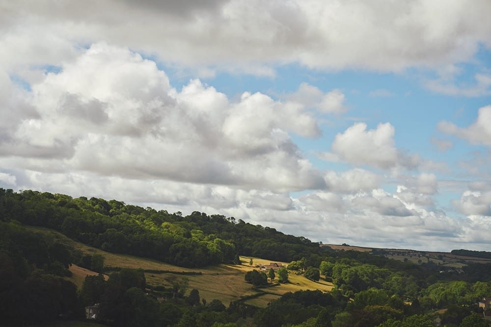 Rolling green hills covered in trees and fields under a partly cloudy blue sky.