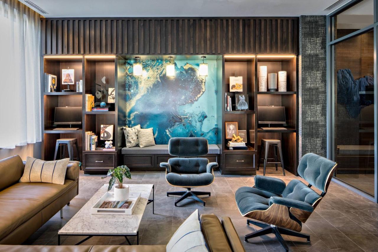 Contemporary hotel lounge with sleek leather sofas, Eames lounge chairs, and dark wood shelving. A large blue-toned abstract wall mural serves as the focal point, framed by built-in bookshelves and soft pendant lighting.