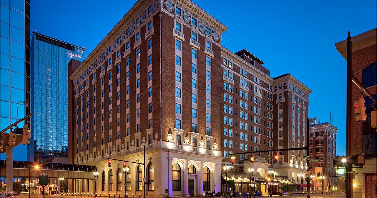 Exterior of the historic Amway Grand Plaza Hotel in Grand Rapids, Michigan, captured at dusk. The grand brick façade is illuminated with architectural lighting, with modern glass towers visible in the background.