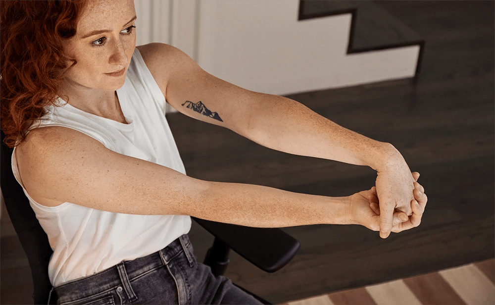 Red-haired woman with freckles stretching her wrists, a mountain tattoo on her arm.