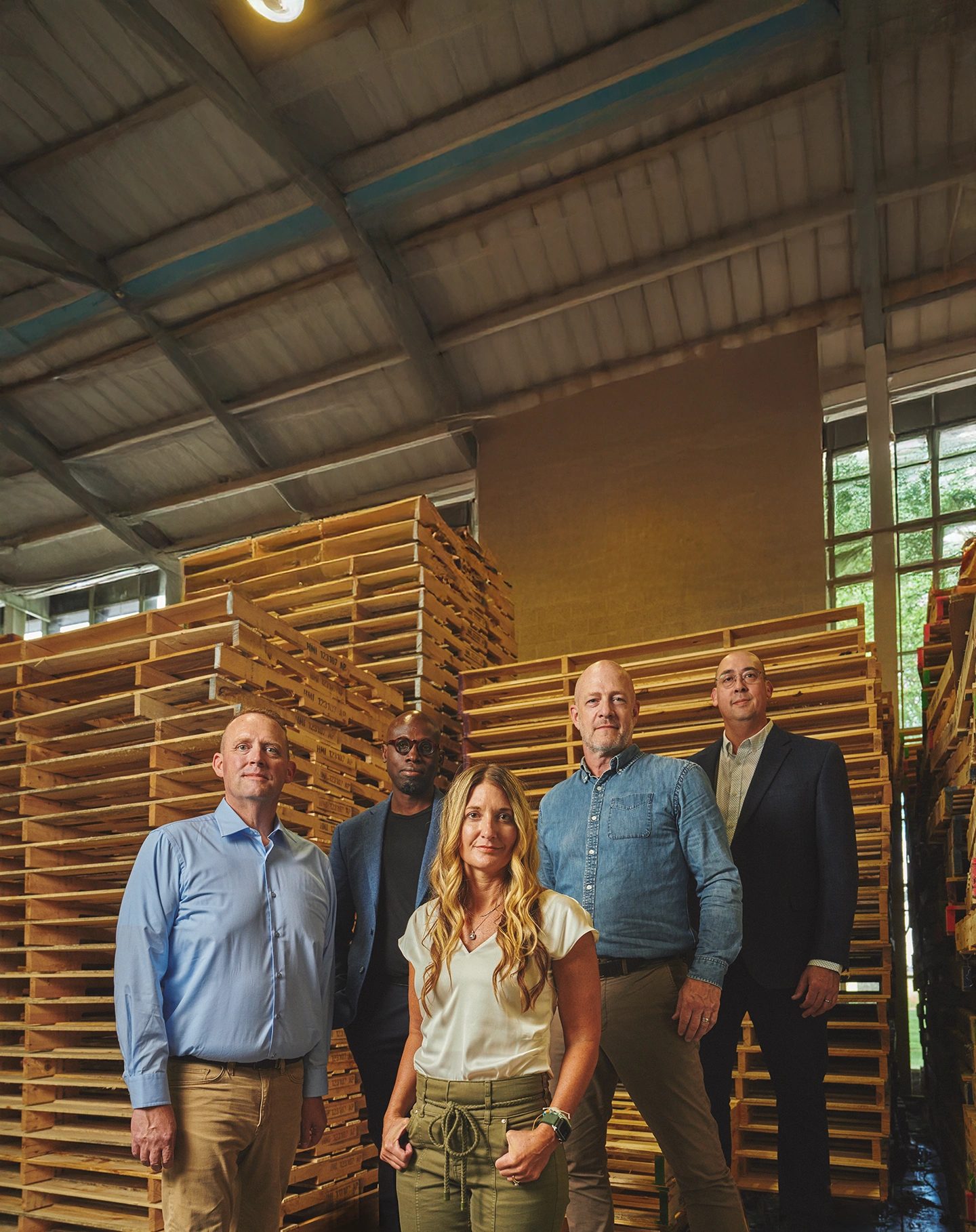 Five people pose in a warehouse with tall stacks of wooden pallets.