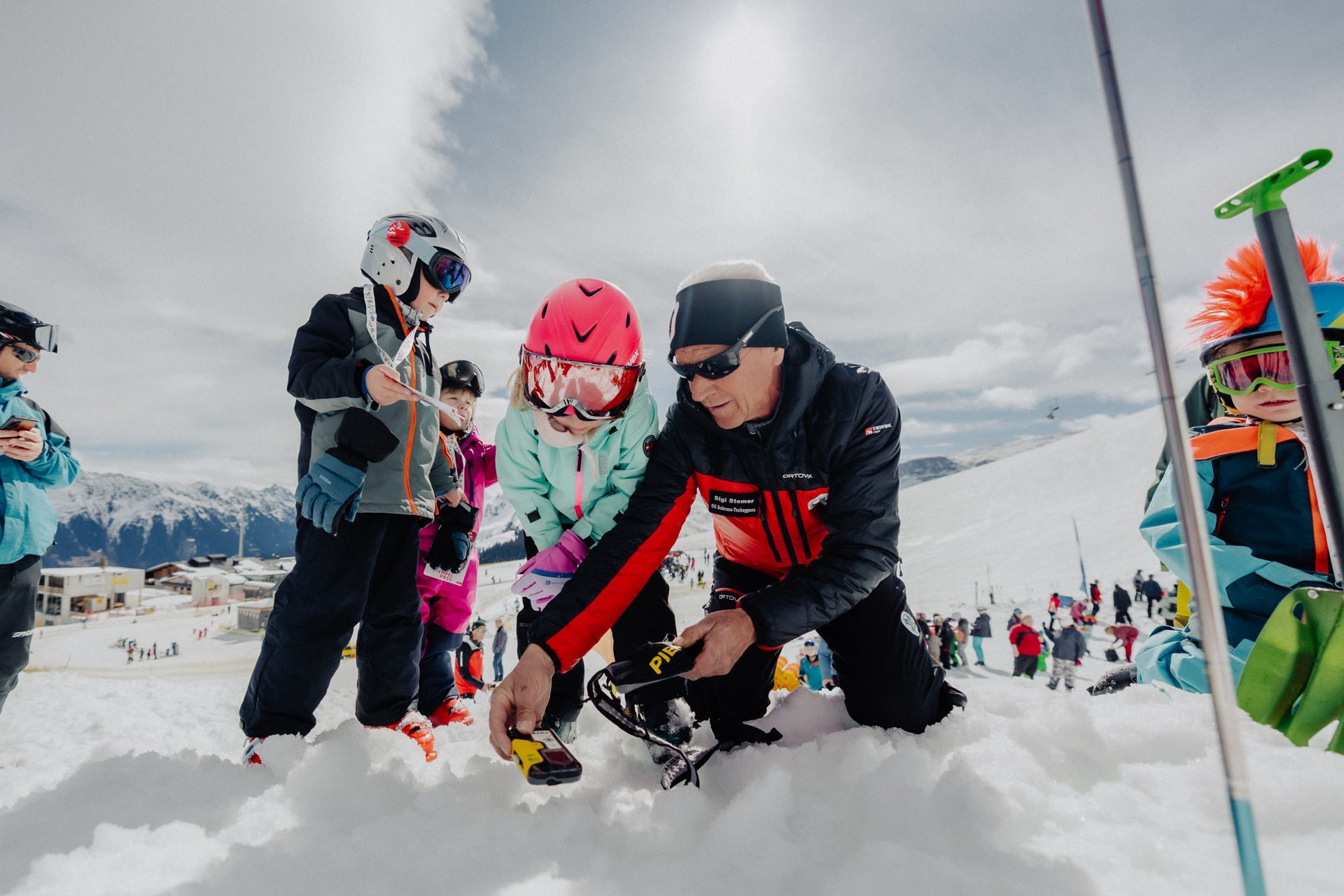 Ski instructor kneeling in the snow, showing equipment to young students wearing helmets and goggles.