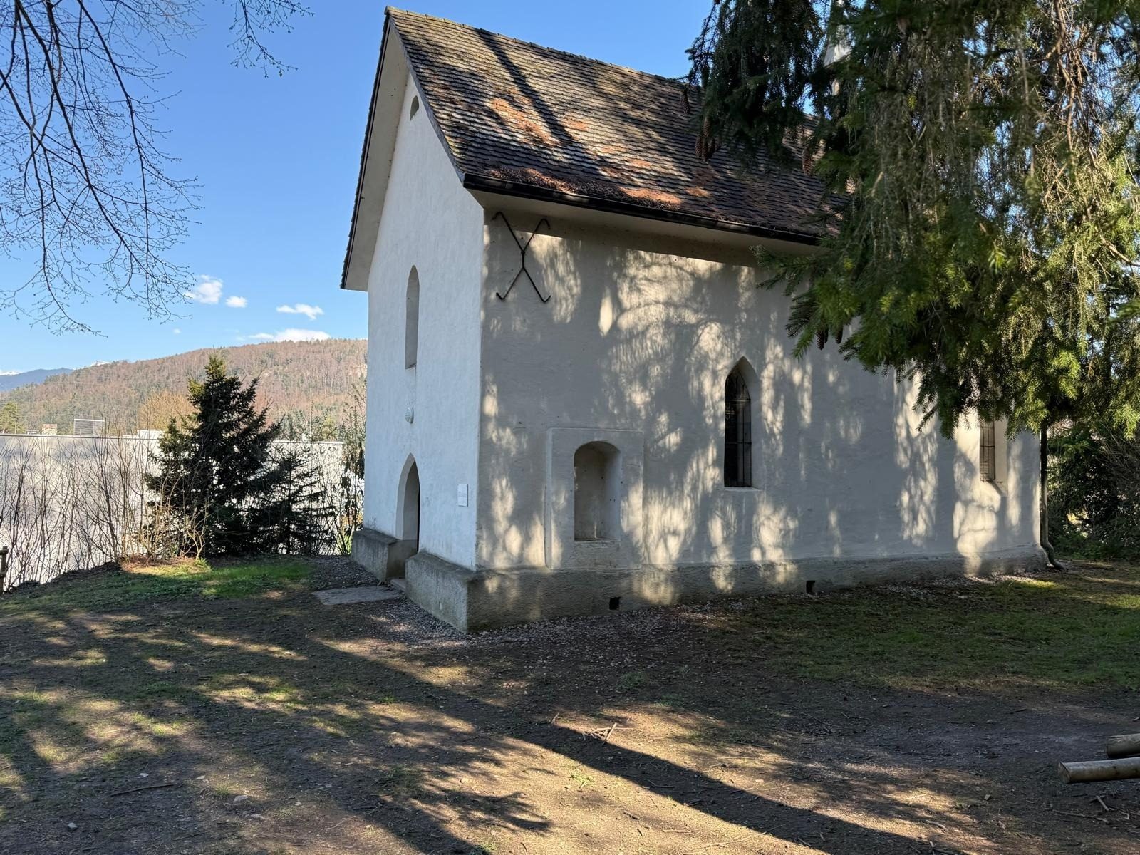 White chapel with brown tiled roof, surrounded by trees and distant mountains under a blue sky, shadows on the ground.