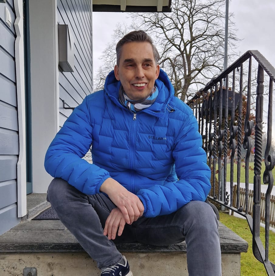 A smiling man in a blue puffer jacket and grey jeans sits on steps outside a house.