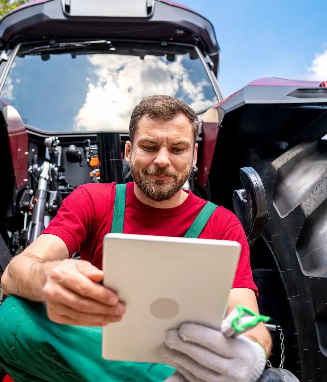 A farmer in overalls and red shirt looks at a tablet while next to a large red tractor.