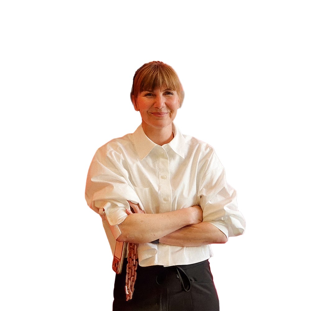 A smiling woman with light brown hair, wearing a white shirt and dark pants, with arms crossed.