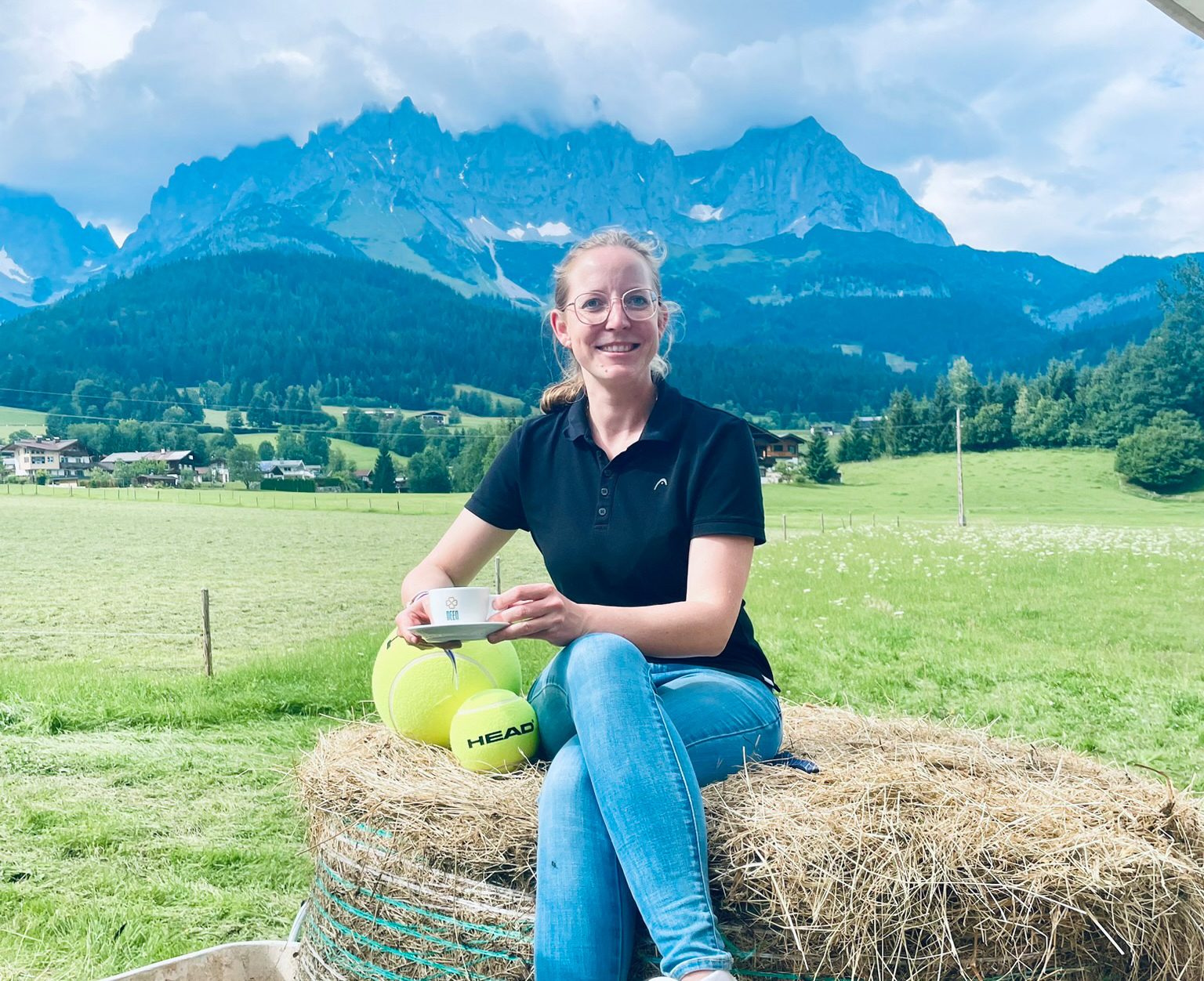 A smiling woman in glasses sits on a hay bale, holding a cup, with tennis balls and mountains.