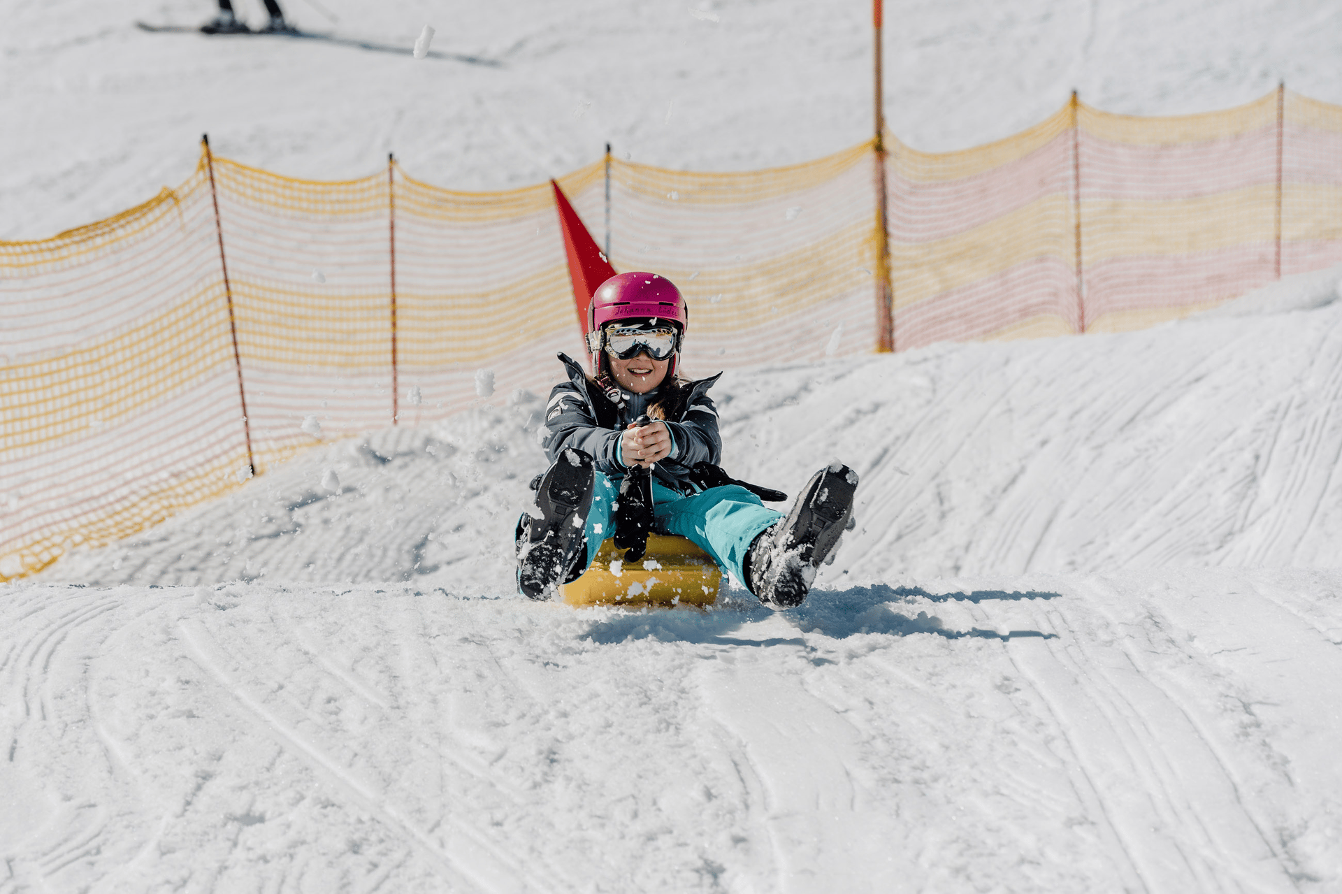 Happy child in a pink helmet and goggles smiling while sledding on a yellow sled down a snowy hill.