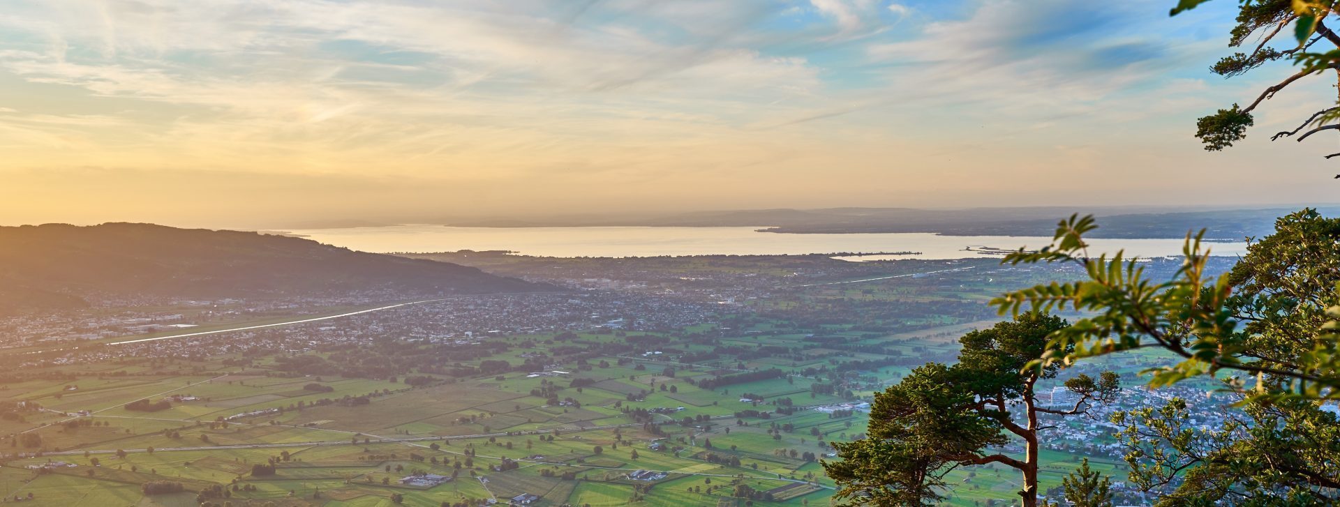 Aerial view of a town, green valley, and lake at sunset, framed by trees.