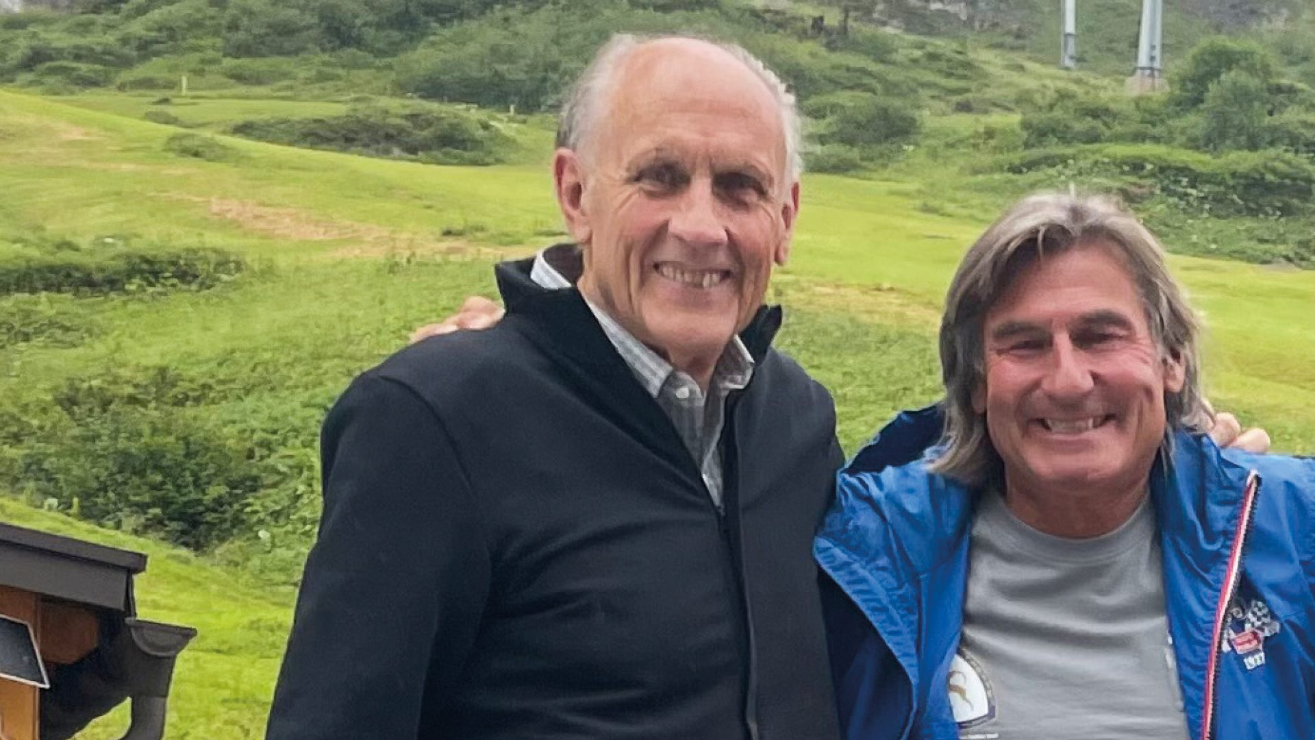 Two smiling men, one older with a dark jacket, the other with long hair and a blue jacket, against green hills.