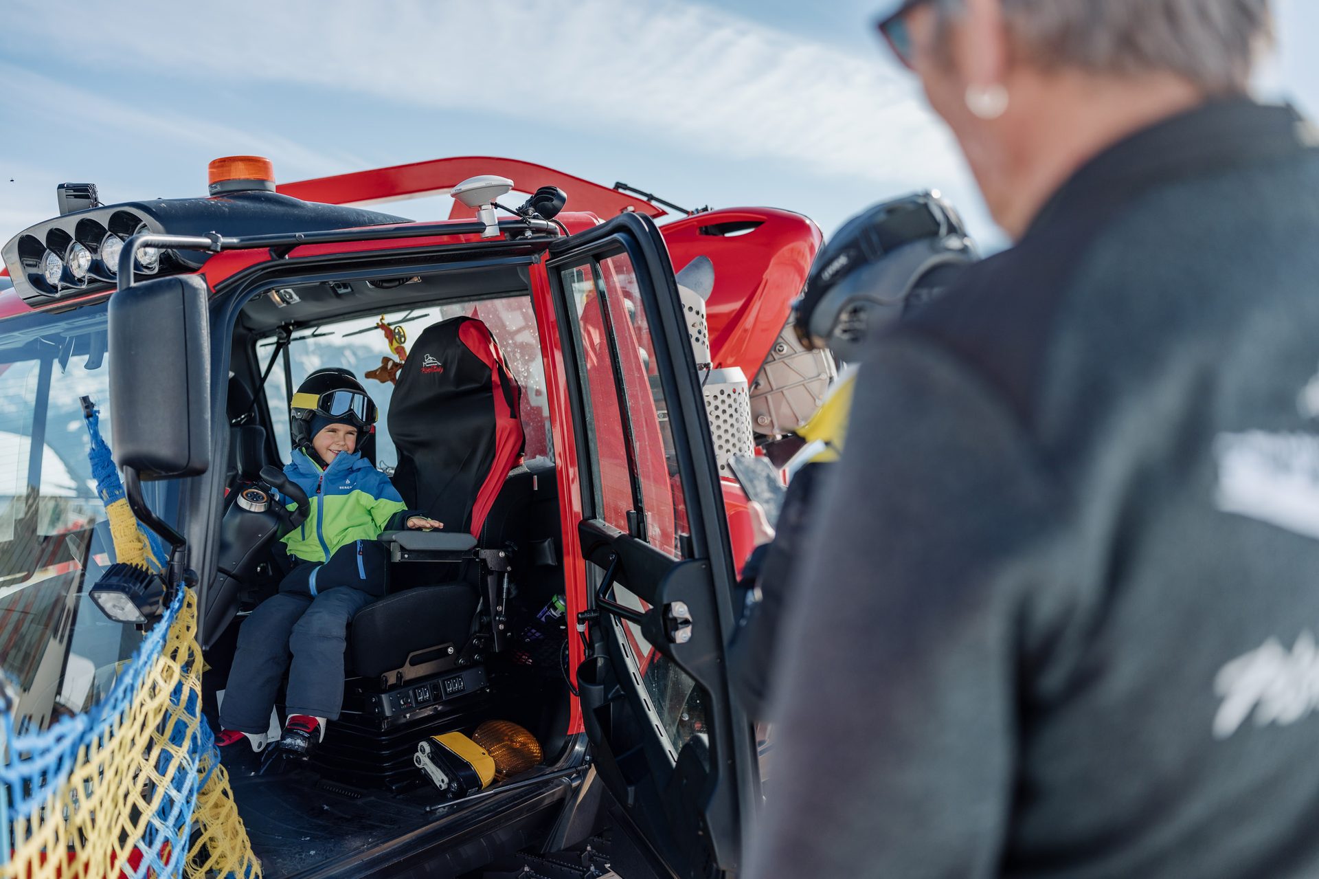 A young child smiling inside the cabin of a red snow groomer, with an adult partially visible.