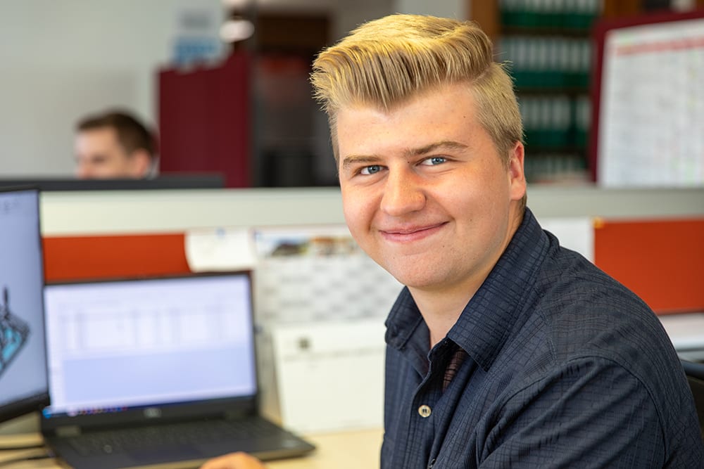 A young, blonde man with blue eyes smiles at the camera in an office setting.