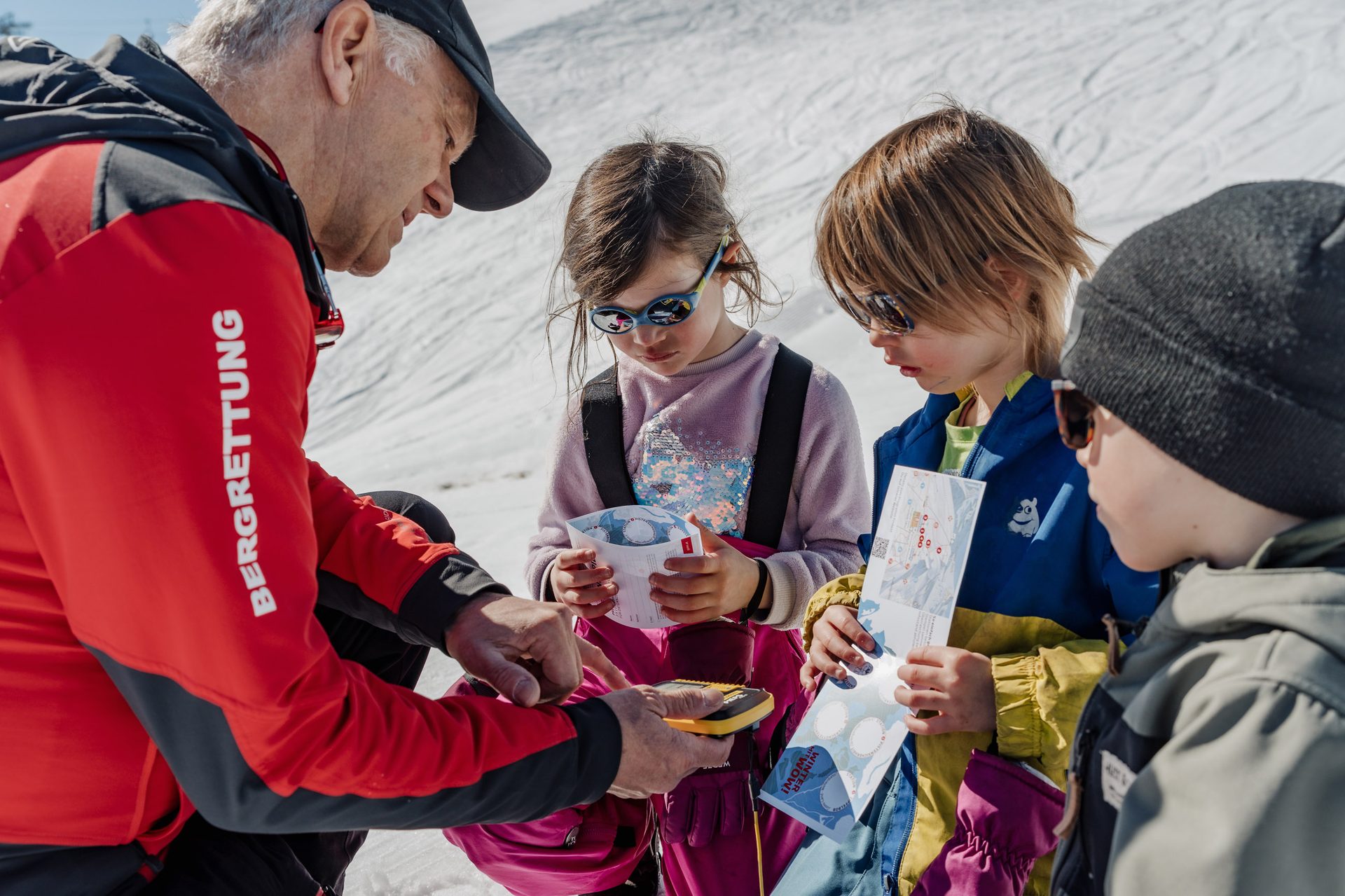 A mountain rescuer teaches three children about maps or avalanche safety in a snowy alpine setting.