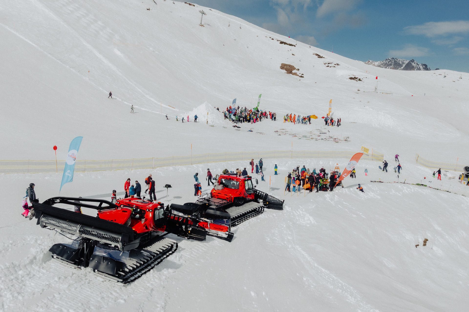 Red snow groomers and many people gathered on a sunny snowy mountain slope.