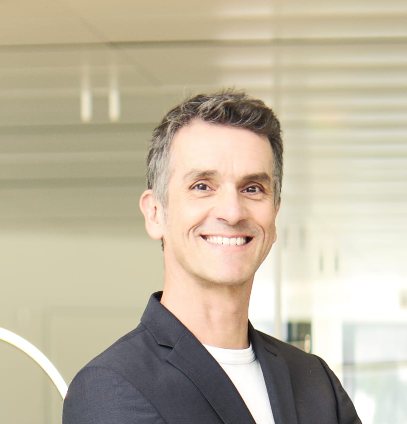 A man with grey hair smiles warmly at the camera, wearing a dark jacket over a white t-shirt.