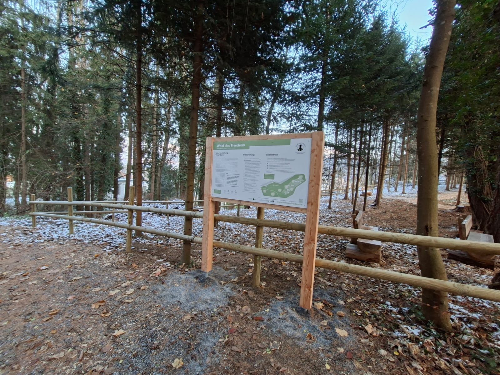 Wooden information sign in a forest with a fence and snow on the ground.