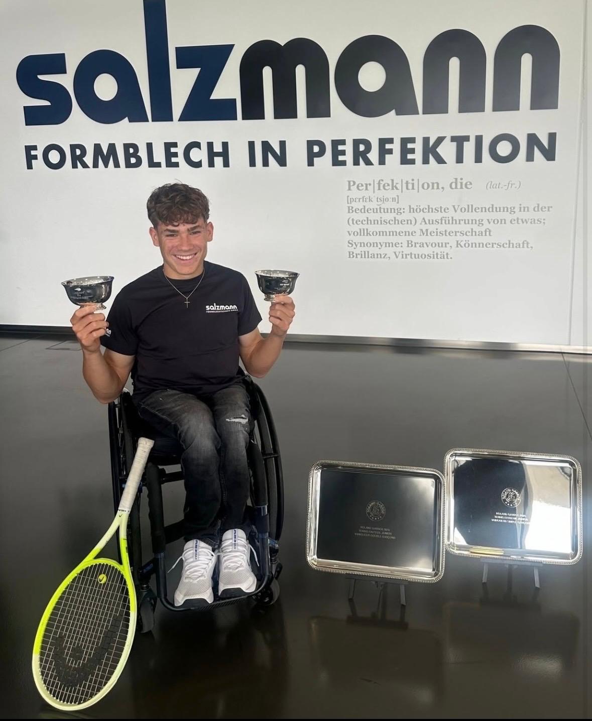 A smiling young man in a wheelchair holds two silver cups, with a tennis racket and two trays nearby.