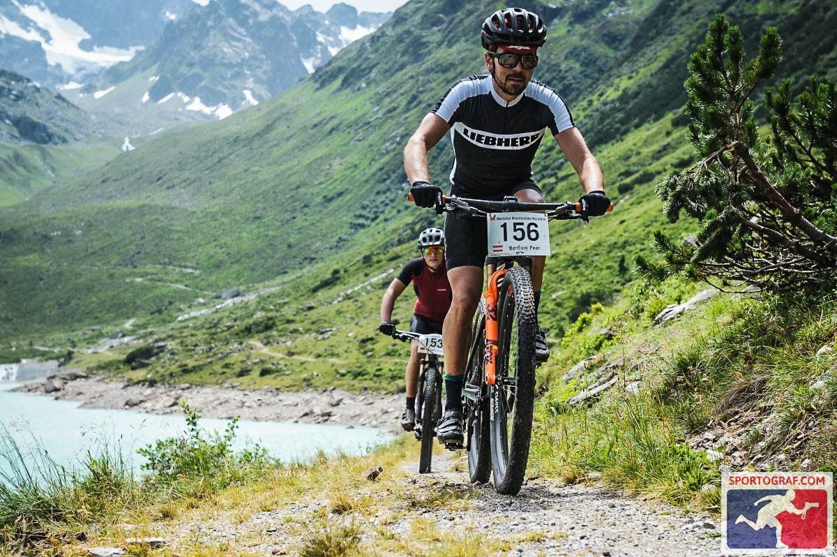 Two mountain bikers on a rocky trail alongside a blue lake with snow-capped mountains in the background.