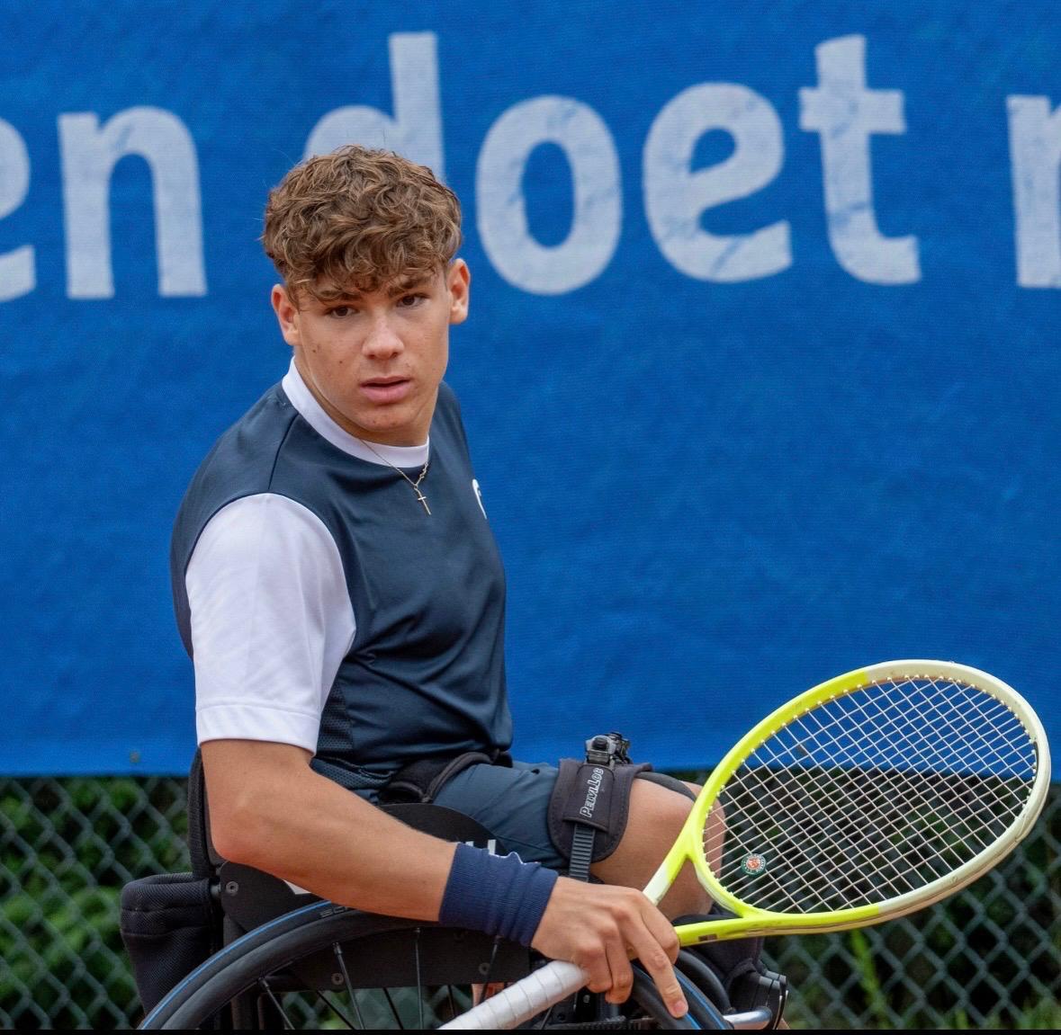 Young male wheelchair tennis player with curly hair holds a racket, looking intently at the viewer.