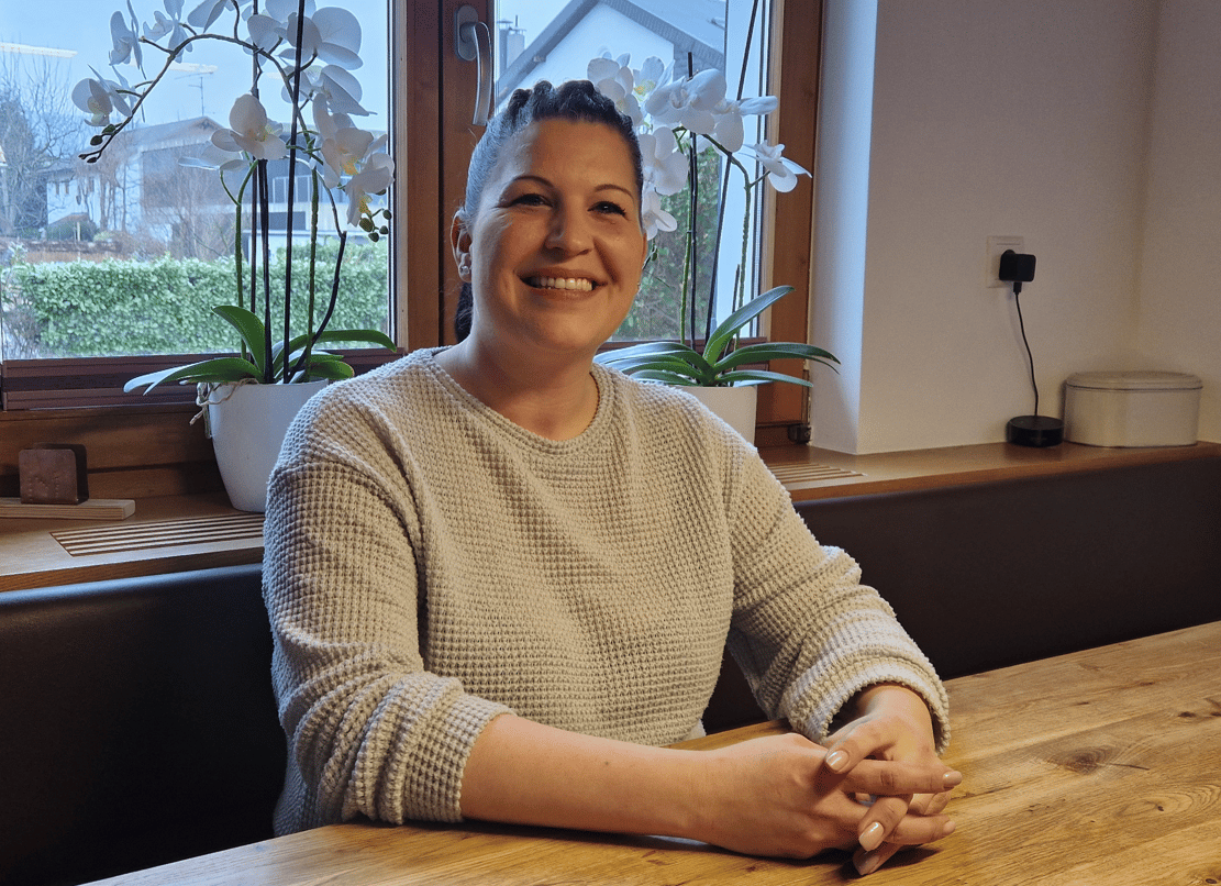 Smiling woman with dark hair in a light grey sweater, seated at a wooden table with orchids behind her.