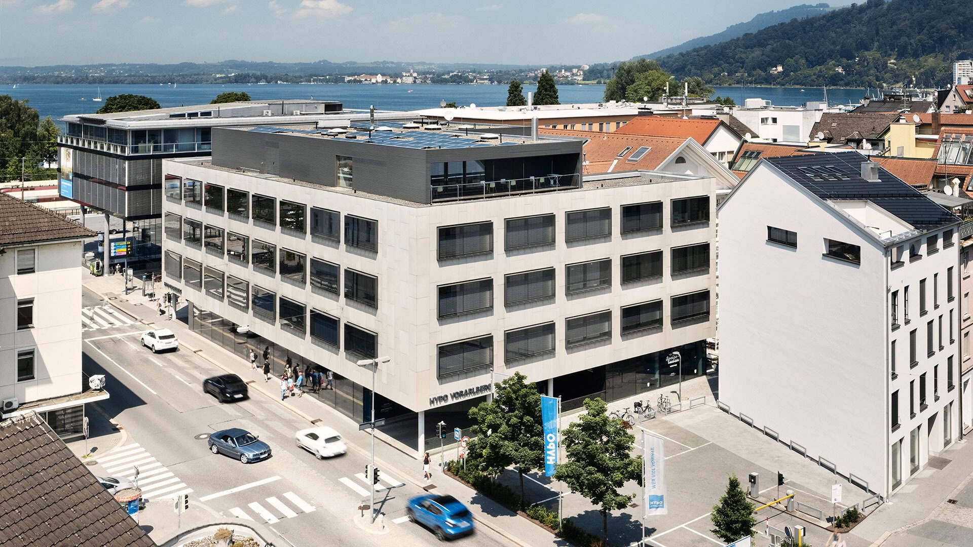 Modern multi-story building with solar panels, street, cars, and a lake with mountains in the background.