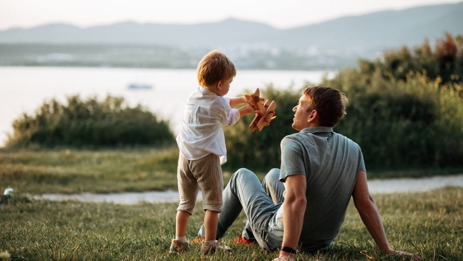 A father and child play with a wooden airplane on grass by a lake.