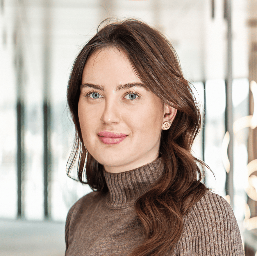 Close-up of a smiling woman with long brown hair and blue eyes, wearing a brown turtleneck.