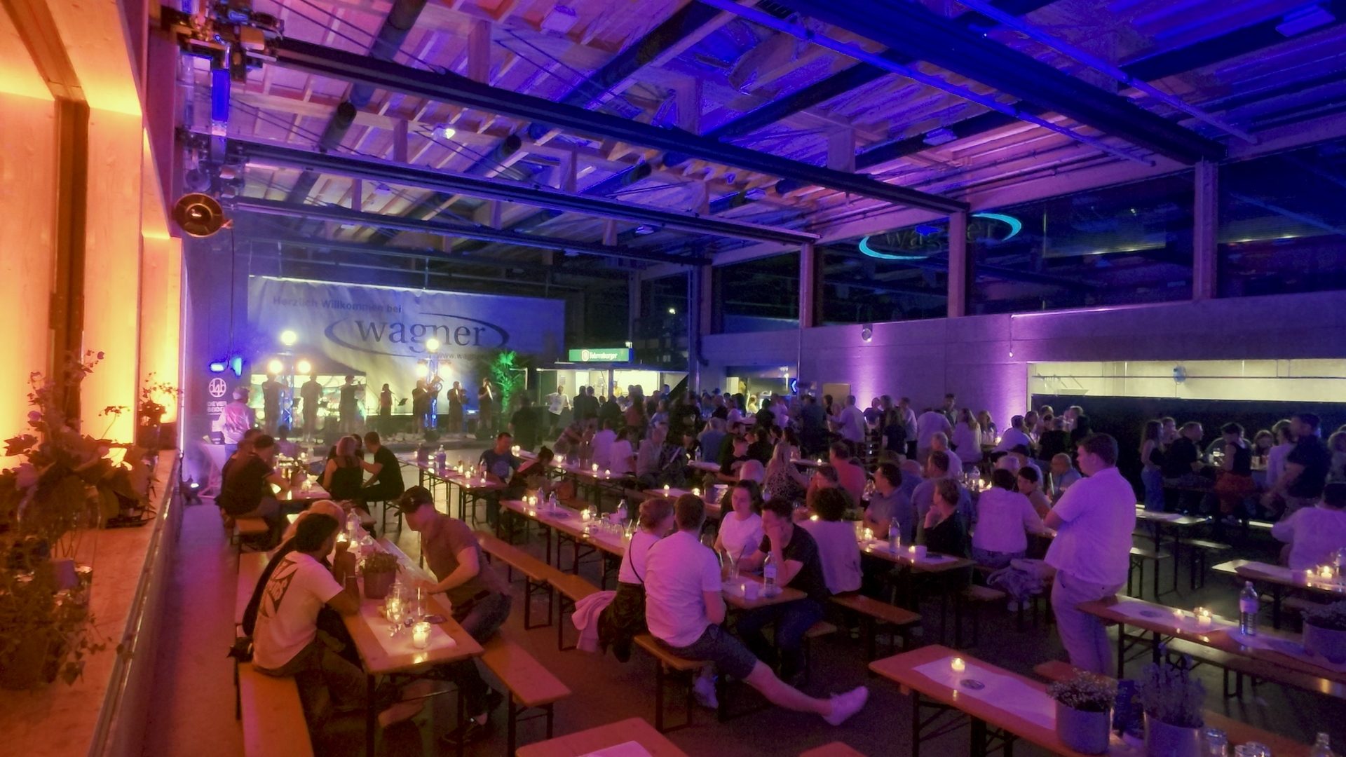 People at a vibrant indoor event with purple and orange lighting, a band performing, and long tables.