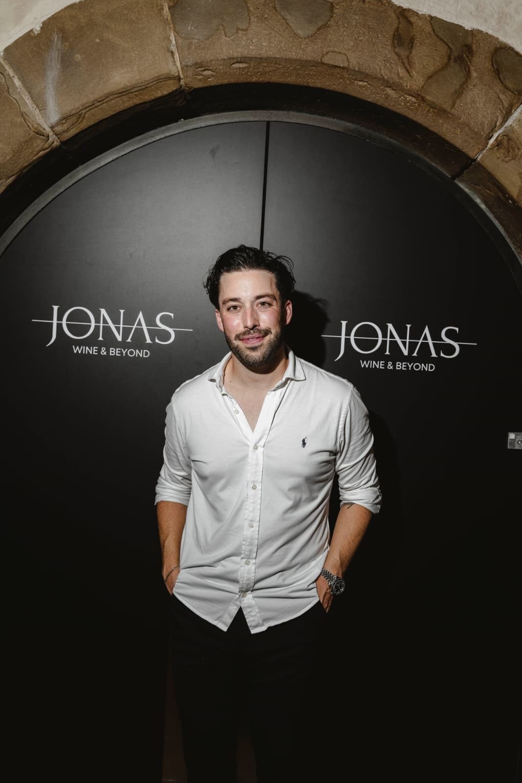 Smiling man in white shirt in front of a dark arched door with "JONAS WINE & BEYOND" text.