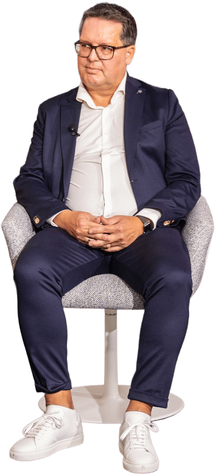 A man in a navy suit, white shirt, and white sneakers sits on a chair, wearing glasses.