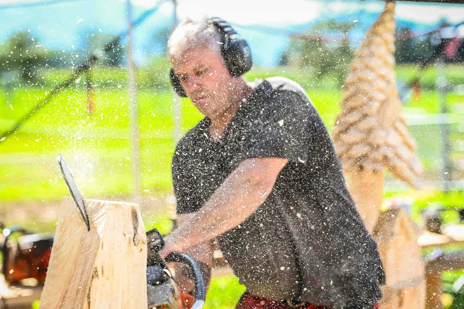 A man in headphones carves wood with a chainsaw, surrounded by sawdust flying in the air.