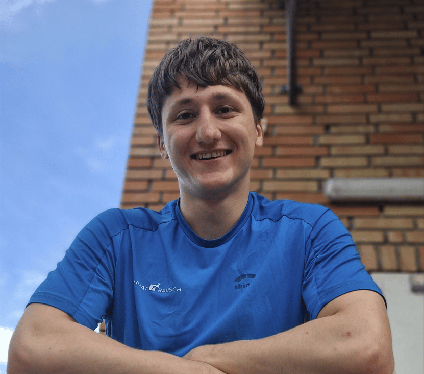 Smiling young man in a blue t-shirt with a brick wall and blue sky in the background.