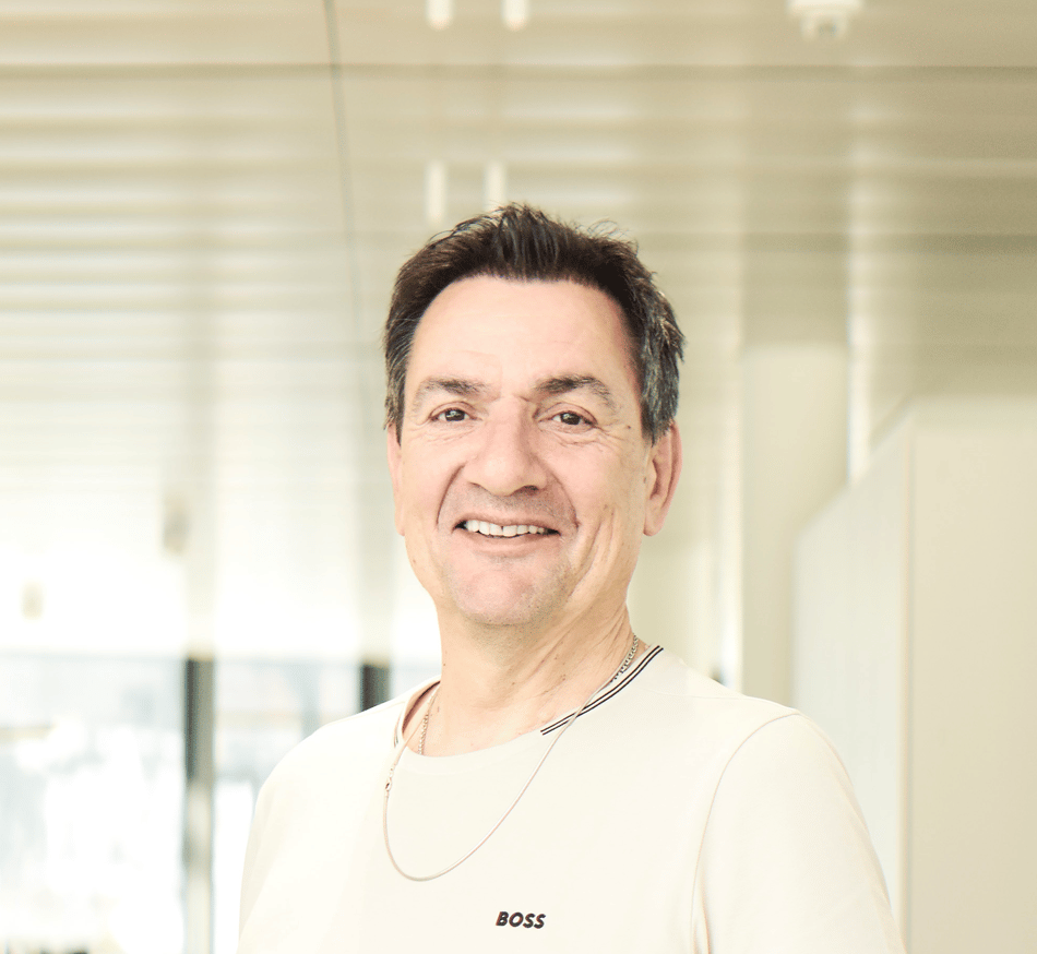 A smiling man in a light t-shirt with a "BOSS" logo and a silver chain looks directly at the camera.