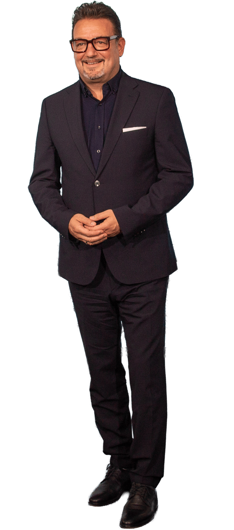 Smiling man in a dark suit and glasses, hands clasped. Full body shot.