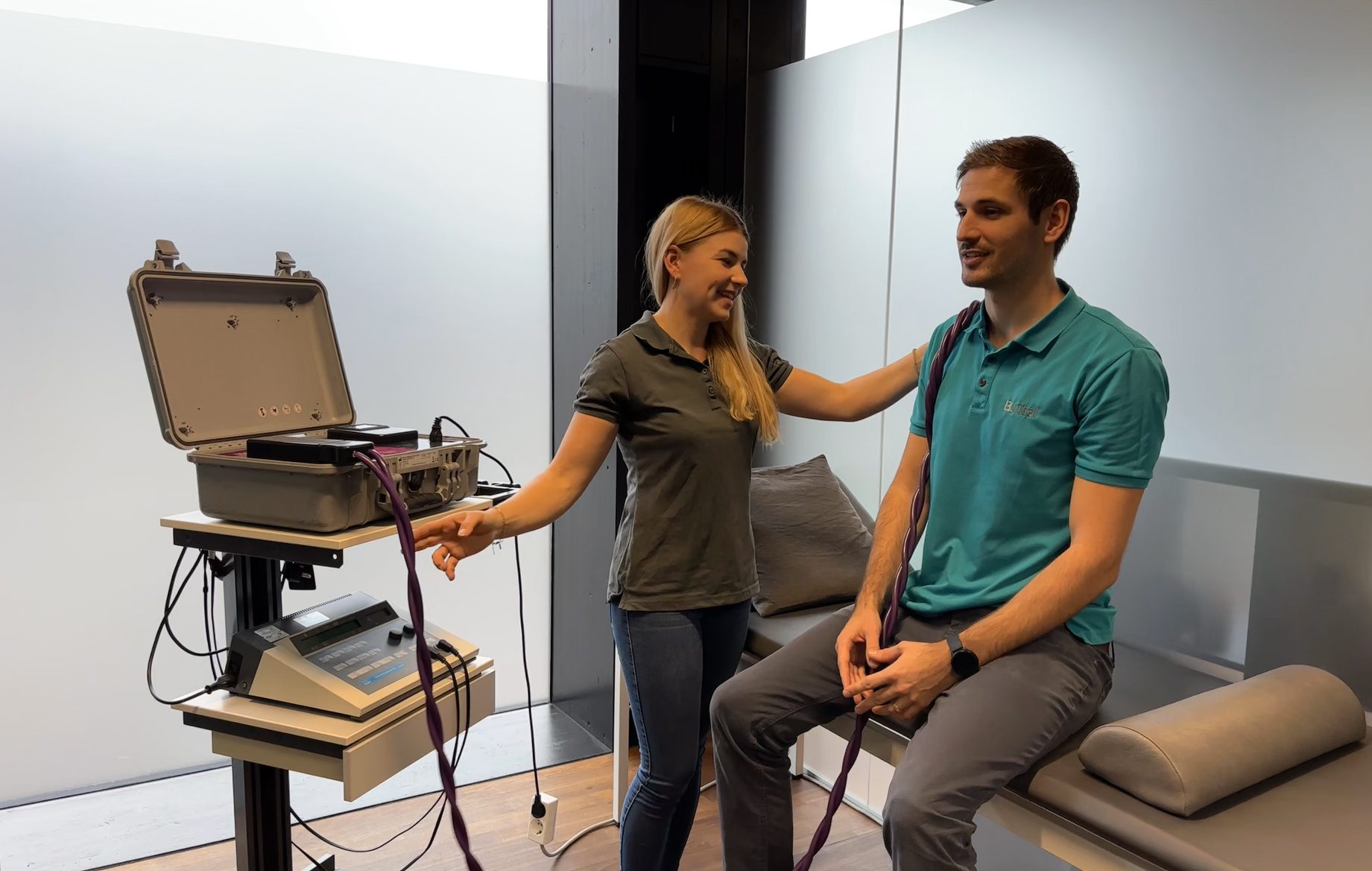 Woman assists man with therapy equipment and cables in a healthcare setting.