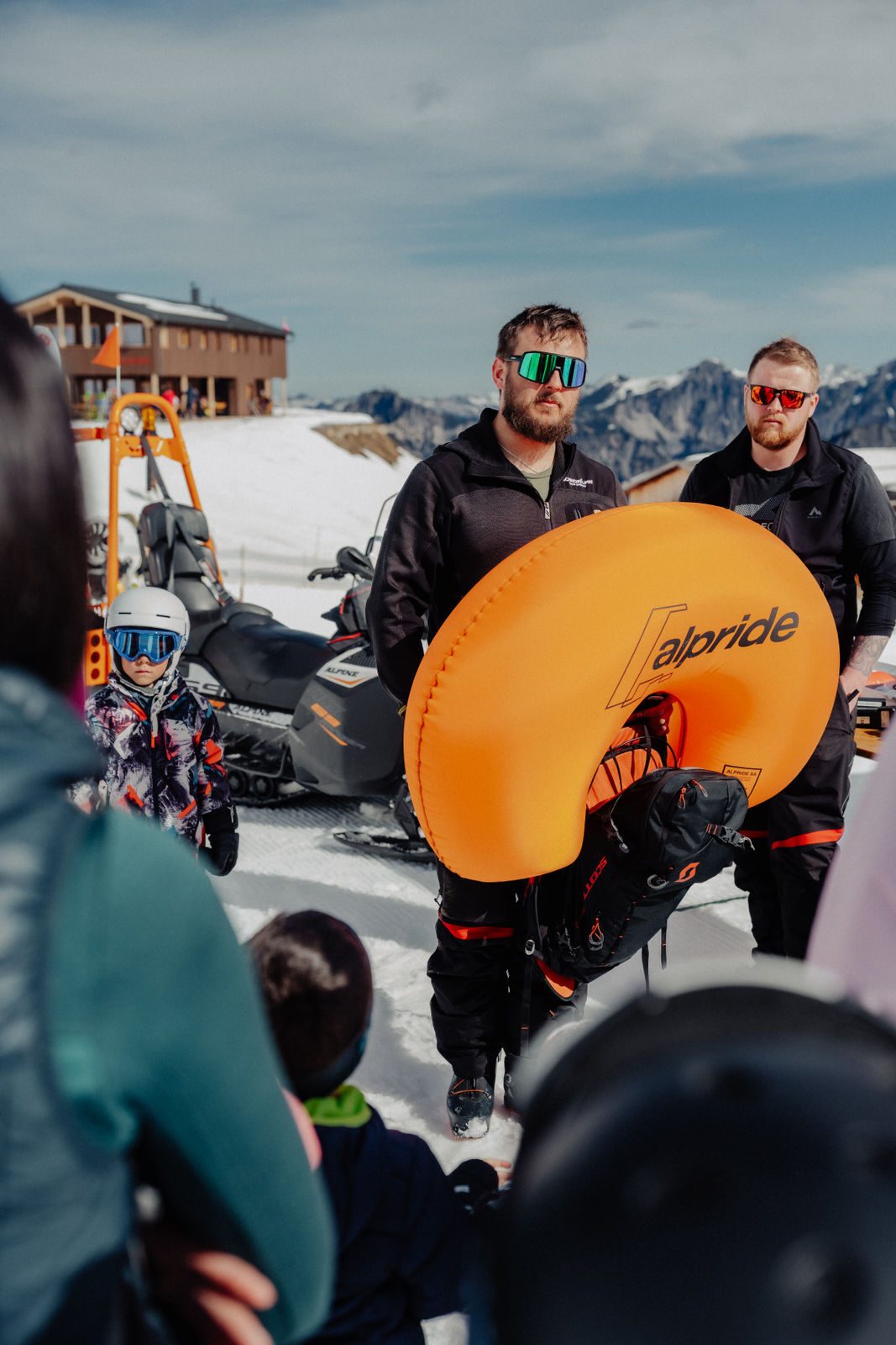 People attending an avalanche safety briefing in the mountains, with an orange airbag demonstration.