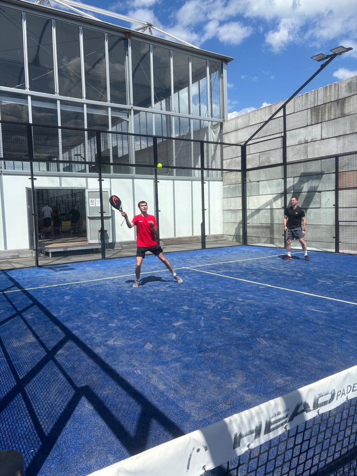 Two men playing padel tennis on an outdoor blue court. A ball is in the air.